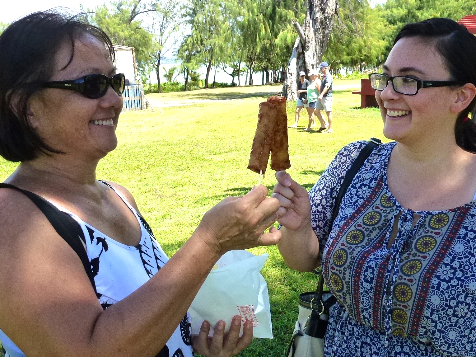 Two women smiling and sharing banana lumpia at the Talofofo Banana Festival in Guam.